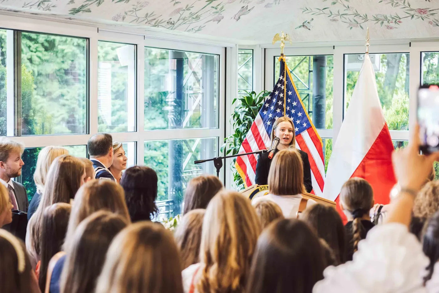 A speaker at the microphone in the Ambassador’s residence, the American and Polish flags behind her and the room watching