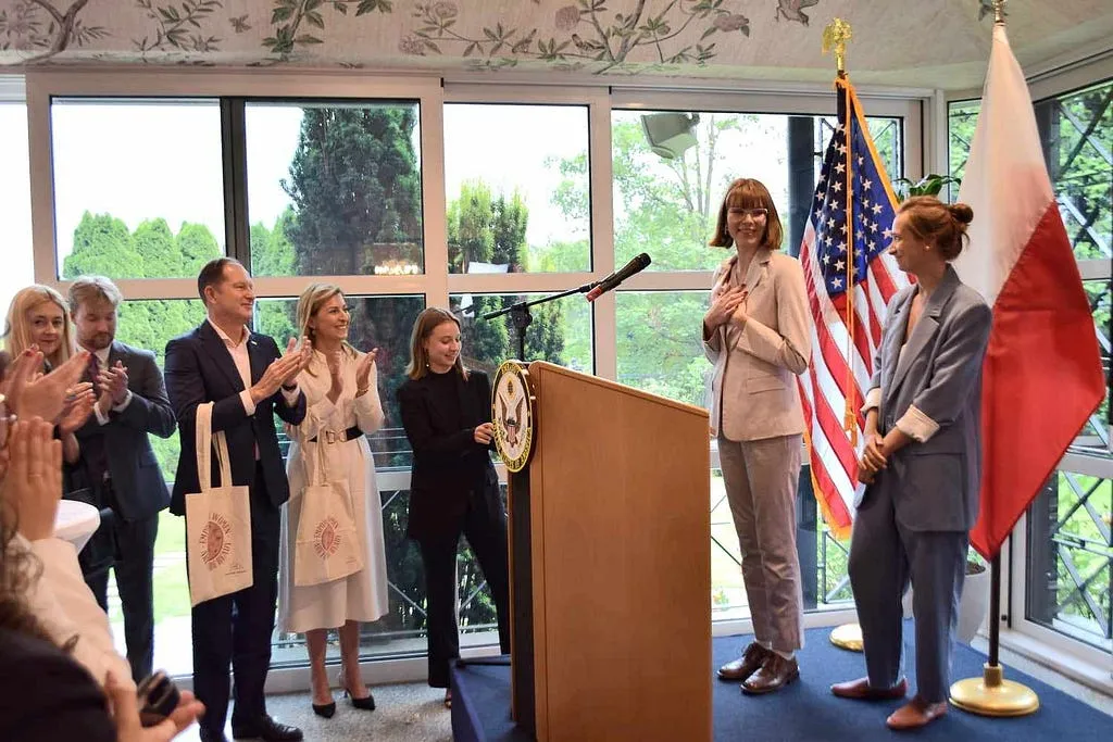 Kalina and others applauding at a lectern between the American and Polish flags, two women standing beside it