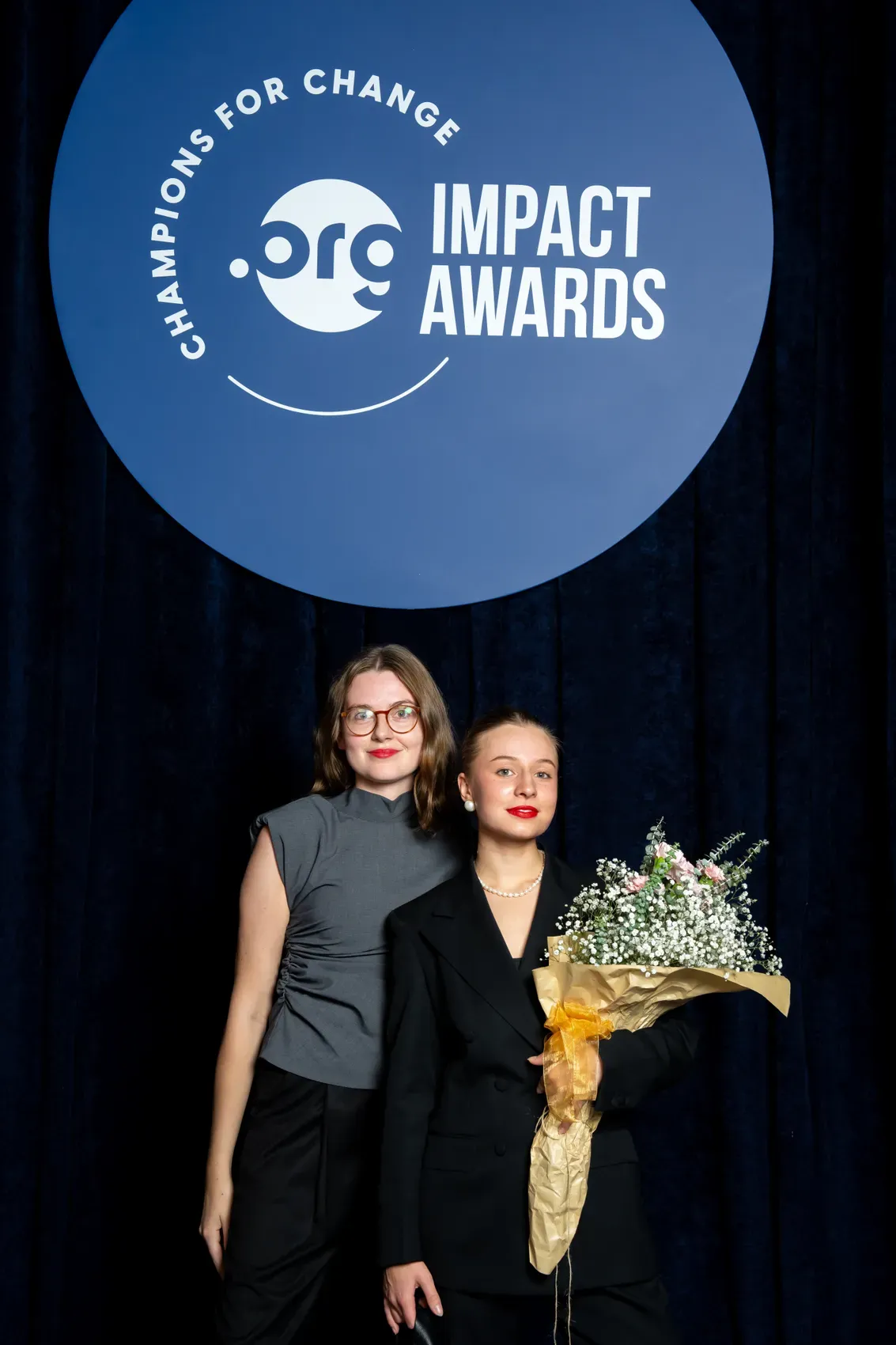 Two of the team on stage at the .ORG Impact Awards, one holding a bouquet, the awards’ roundel lit behind them