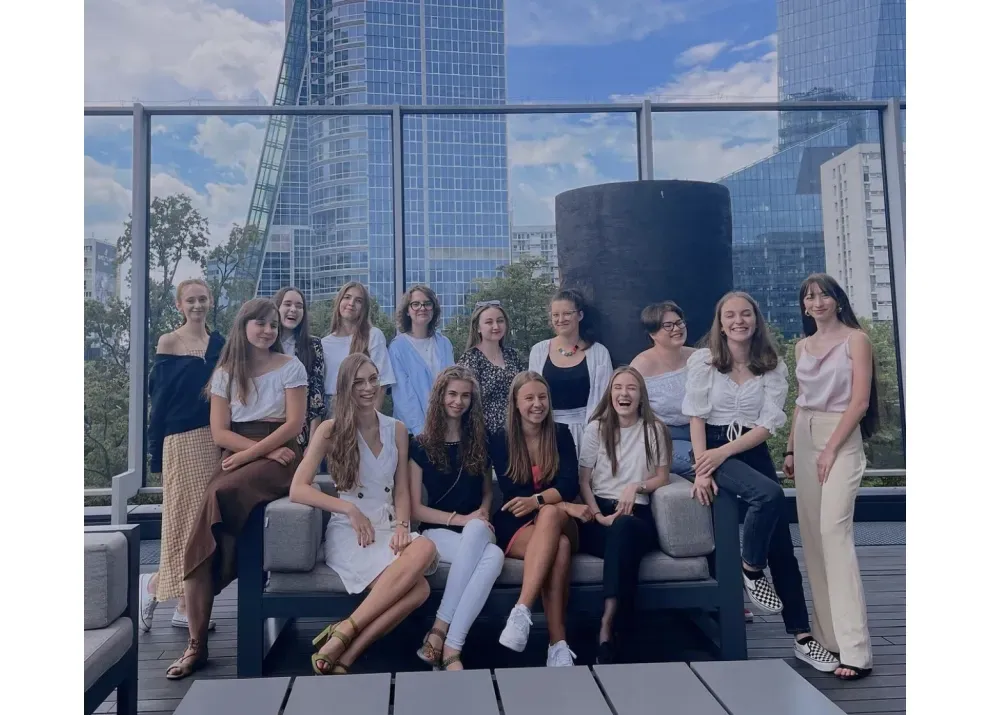 A dozen young women sitting together on a rooftop terrace, city towers behind them